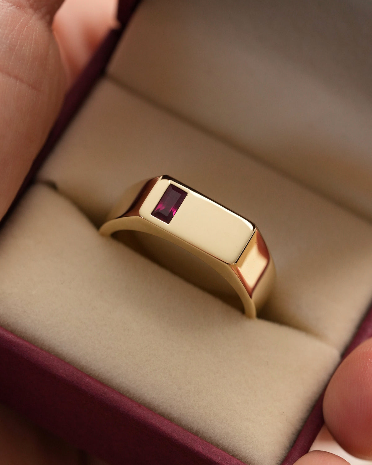 A polished yellow gold signet ring with a rectangular dark red gemstone, displayed inside a cream-colored jewelry box held by hands.