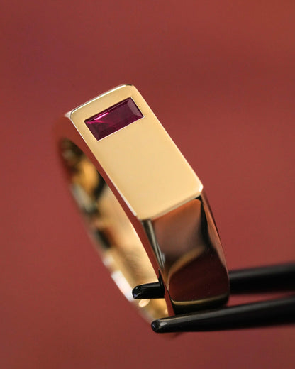 A close-up of a gold ring with a rectangular bezel set with a small rectangular purple gemstone, held by black tweezers against a warm reddish-brown background.