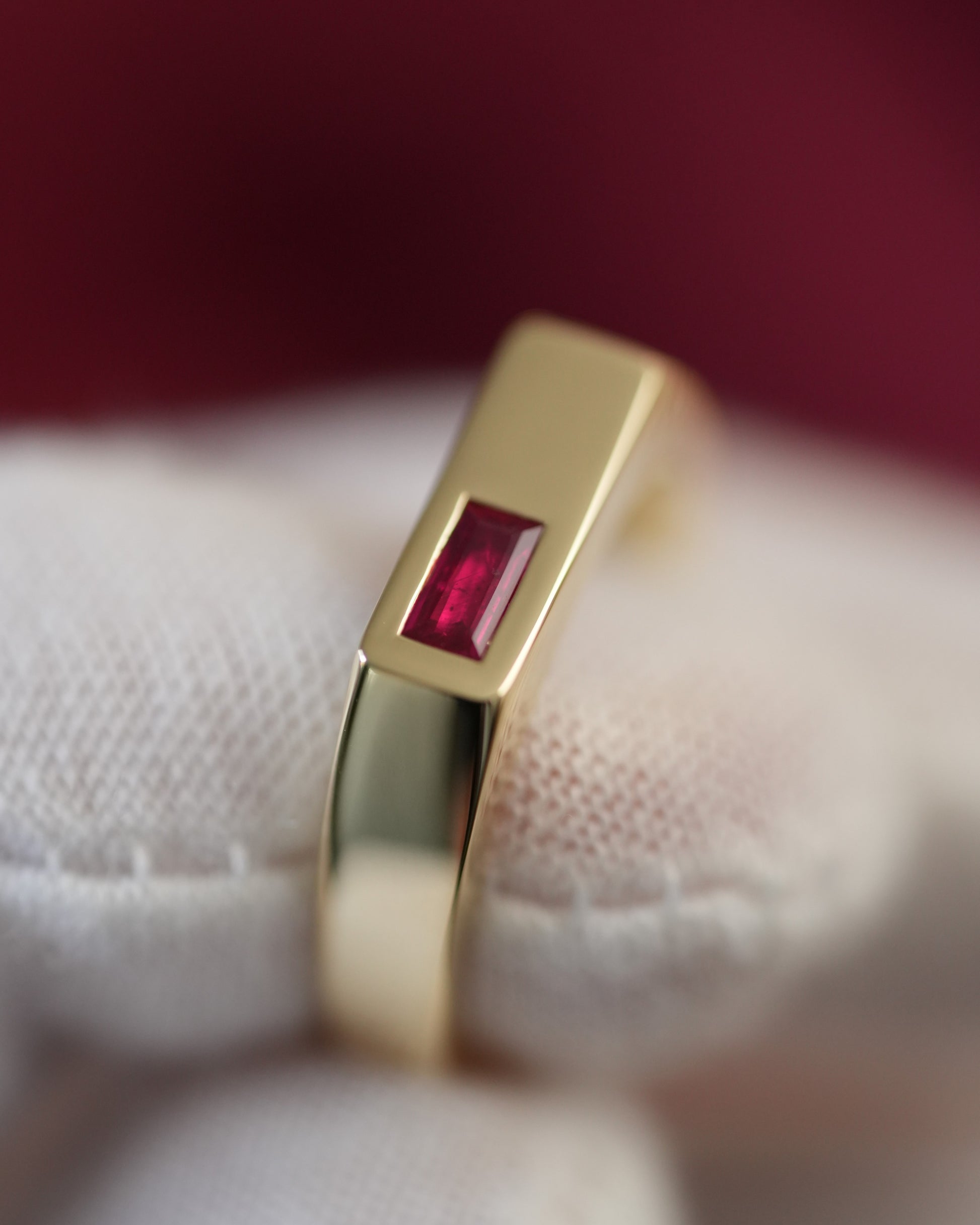 A gold ring with a rectangular red gemstone, displayed on a white textured fabric surface against a dark red background.